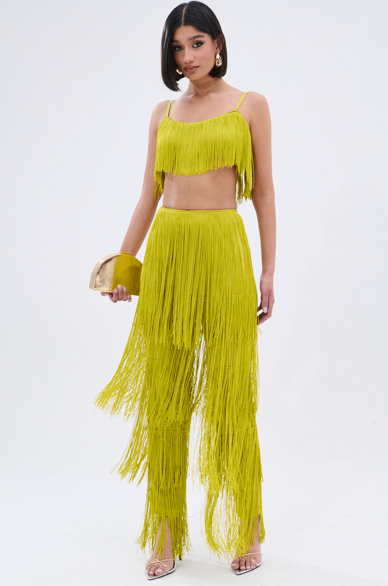 A woman wears the OH SO FRINGE PANTS IN GREEN—stretchy, high-rise, two-piece fringe pants with a matching crop top—holding a gold clutch and standing in clear high heels against a plain white background.