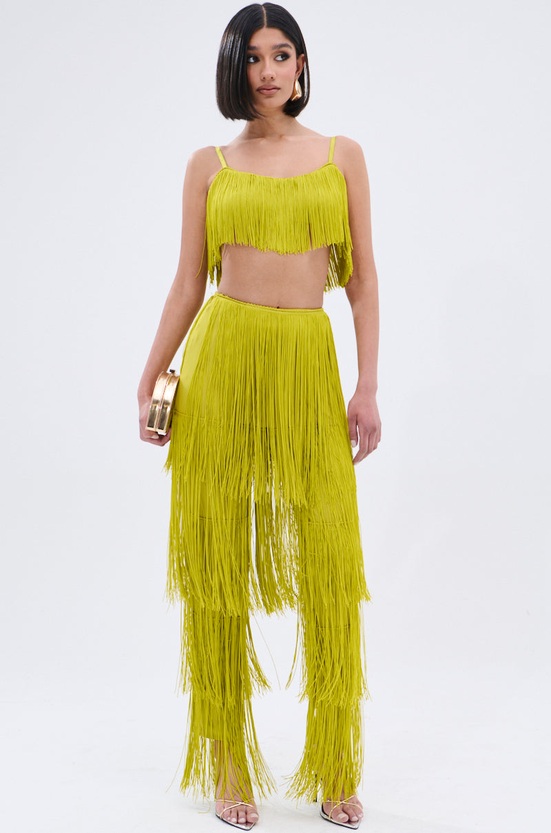 A woman stands against a white background wearing OH SO FRINGE PANTS IN GREEN with a matching green fringe crop top, holding a small gold clutch and wearing open-toed heels.