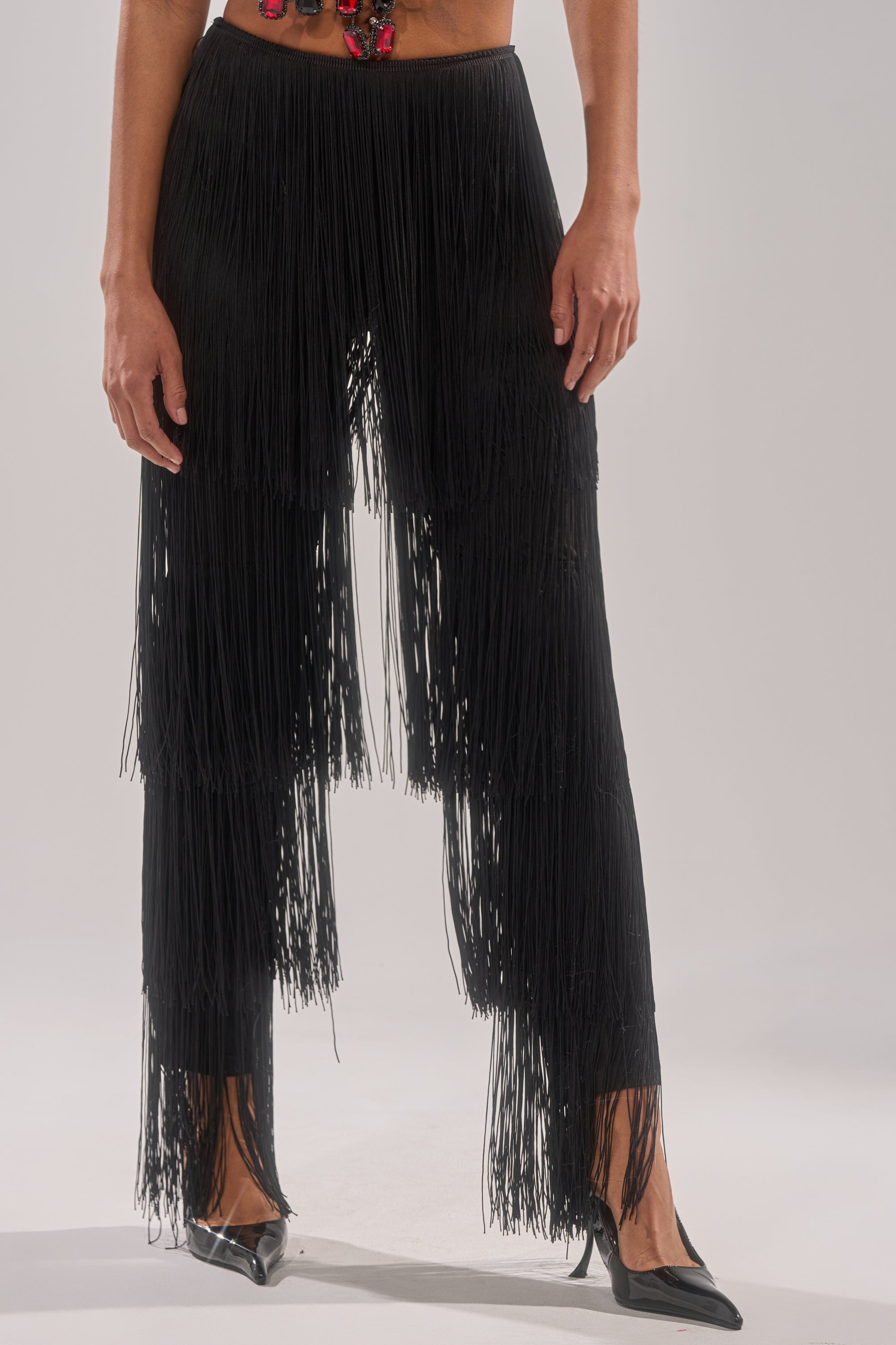 A person stands against a plain light background wearing the OH SO FRINGE PANTS IN BLACK and black pointed-toe heels, hands relaxed by their sides.