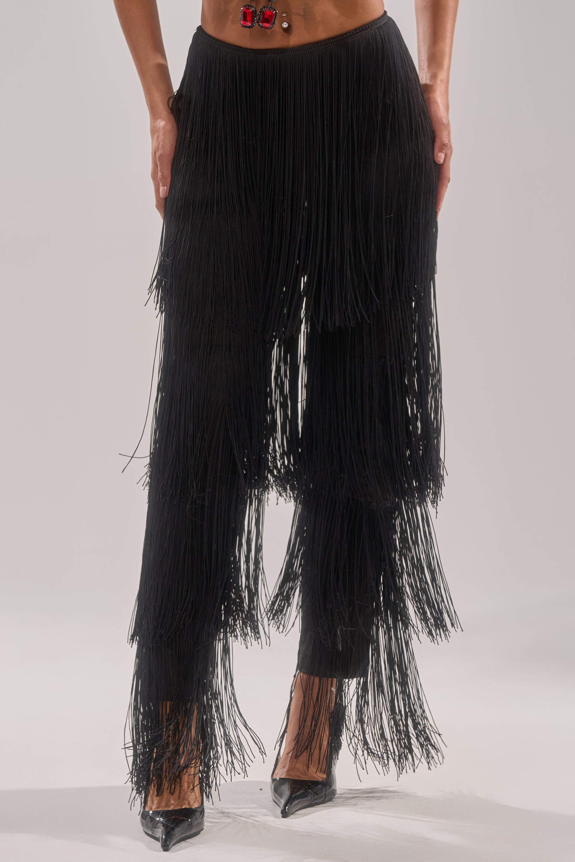 A person stands with hands on hips in OH SO FRINGE PANTS IN BLACK, featuring multiple layers of long, black fringe and tassel details. They pair the high-rise pants with black pointed-toe heels against a plain light background.