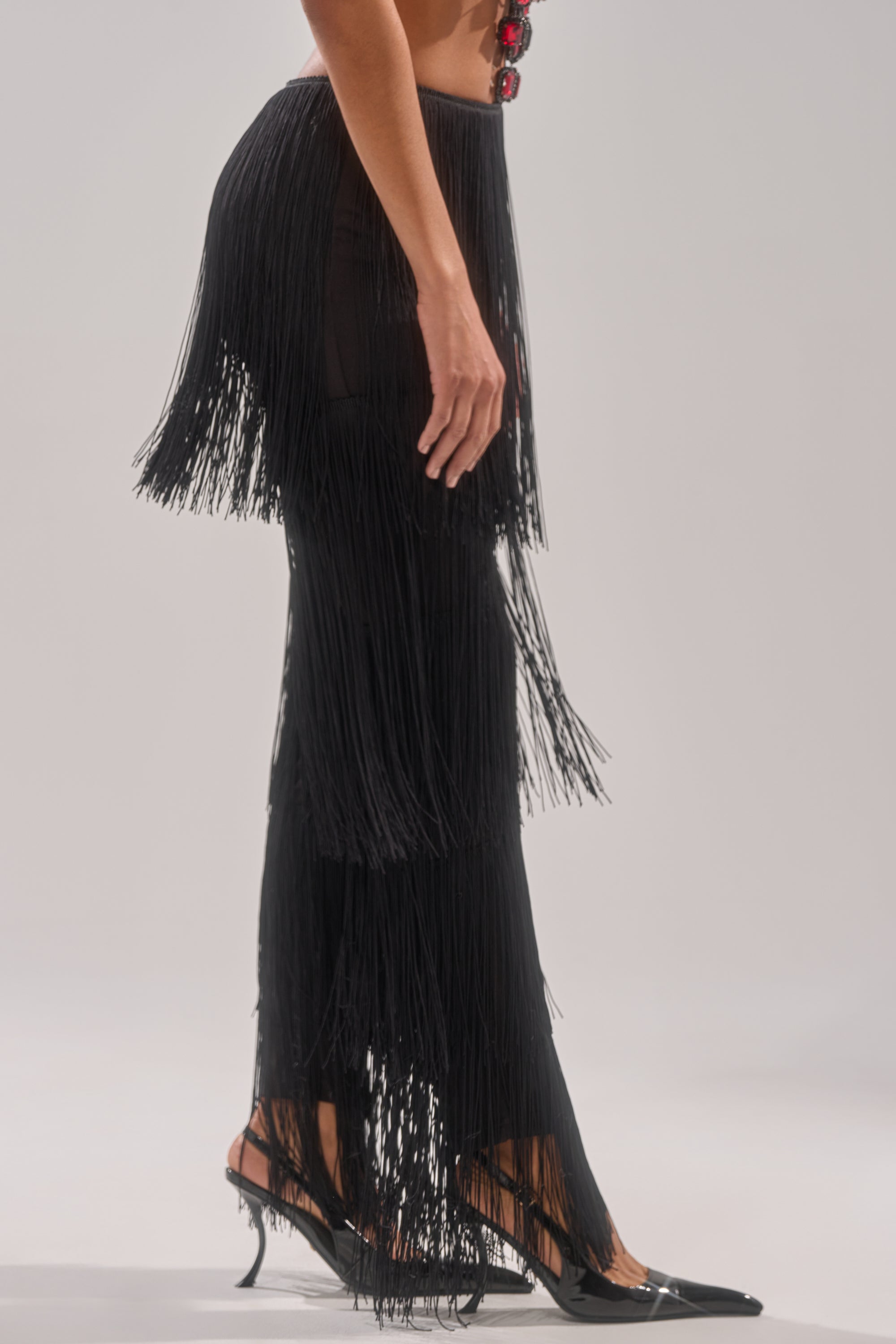 A person models the OH SO FRINGE PANTS IN BLACK—multi-layered long fringe sways with movement. Styled with black pointed-toe heels, they stand against a plain light background.