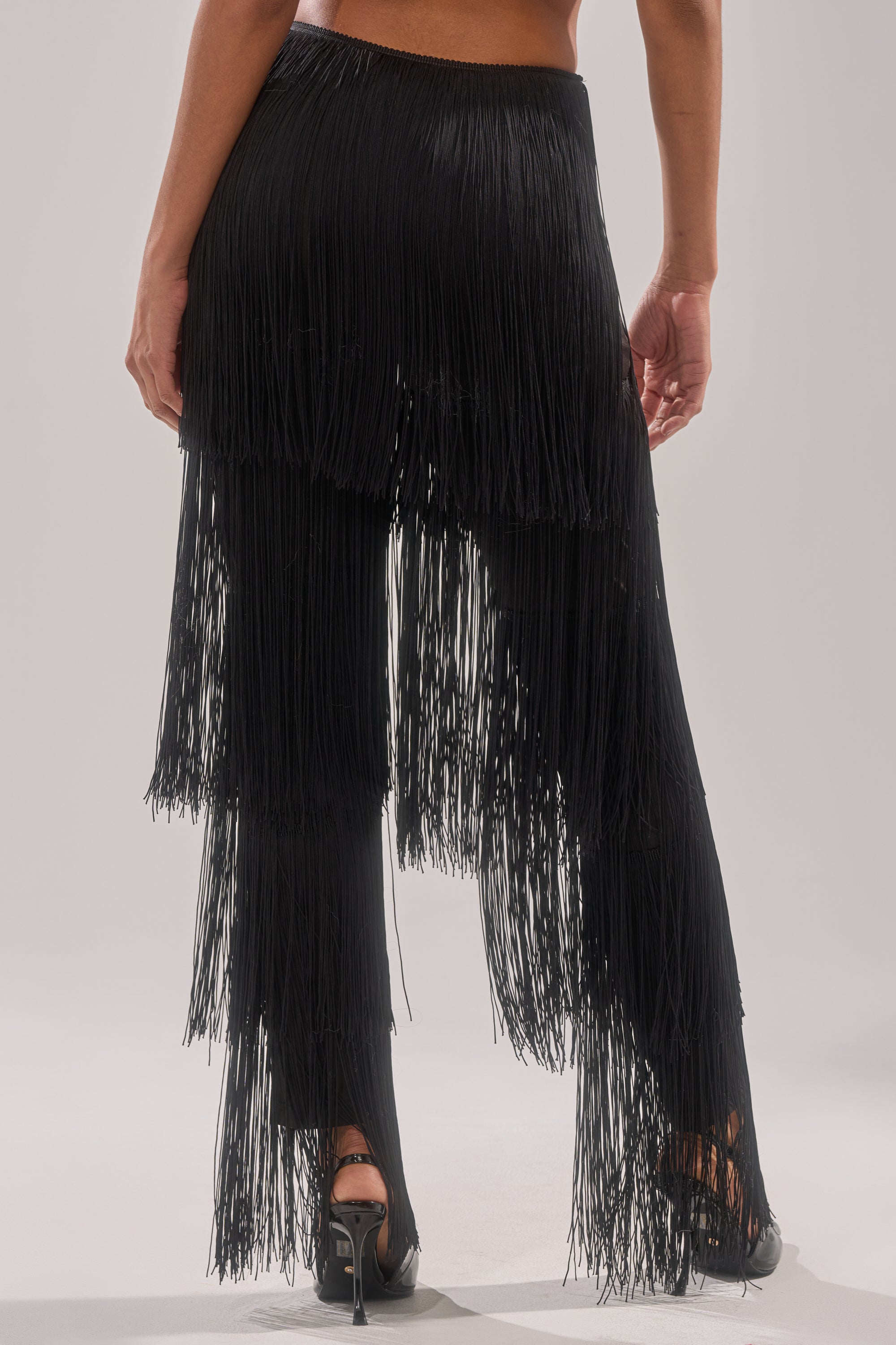 A person wearing high-heeled shoes and the OH SO FRINGE PANTS IN BLACK, seen from behind. The pants feature layered, long fringe detailing cascading down the legs.