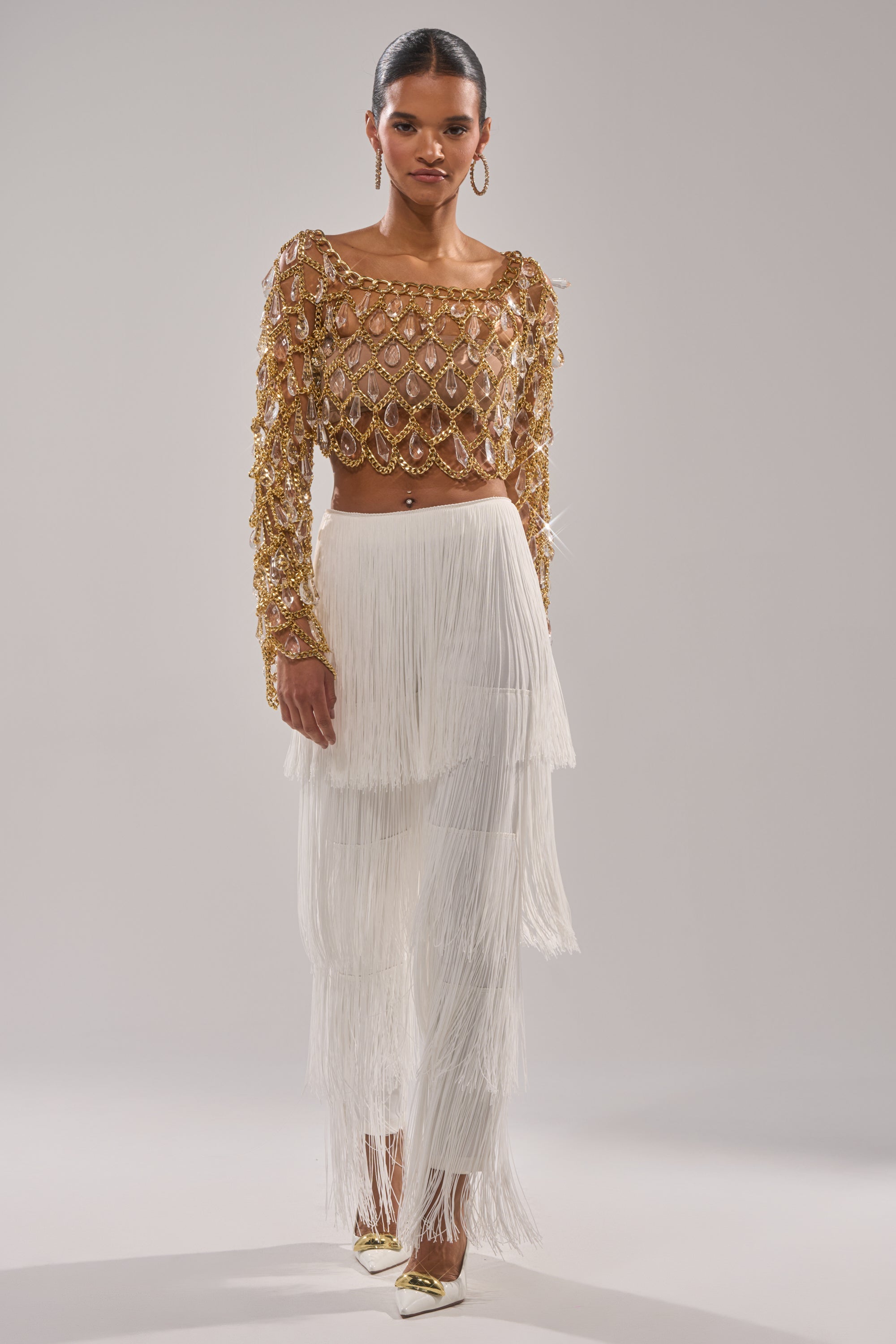 A woman poses against a plain light background wearing the OH SO FRINGE PANTS IN WHITE, styled with a gold embellished long-sleeve crop top, hoop earrings, gold pointed heels, and her hair pulled back. 0
