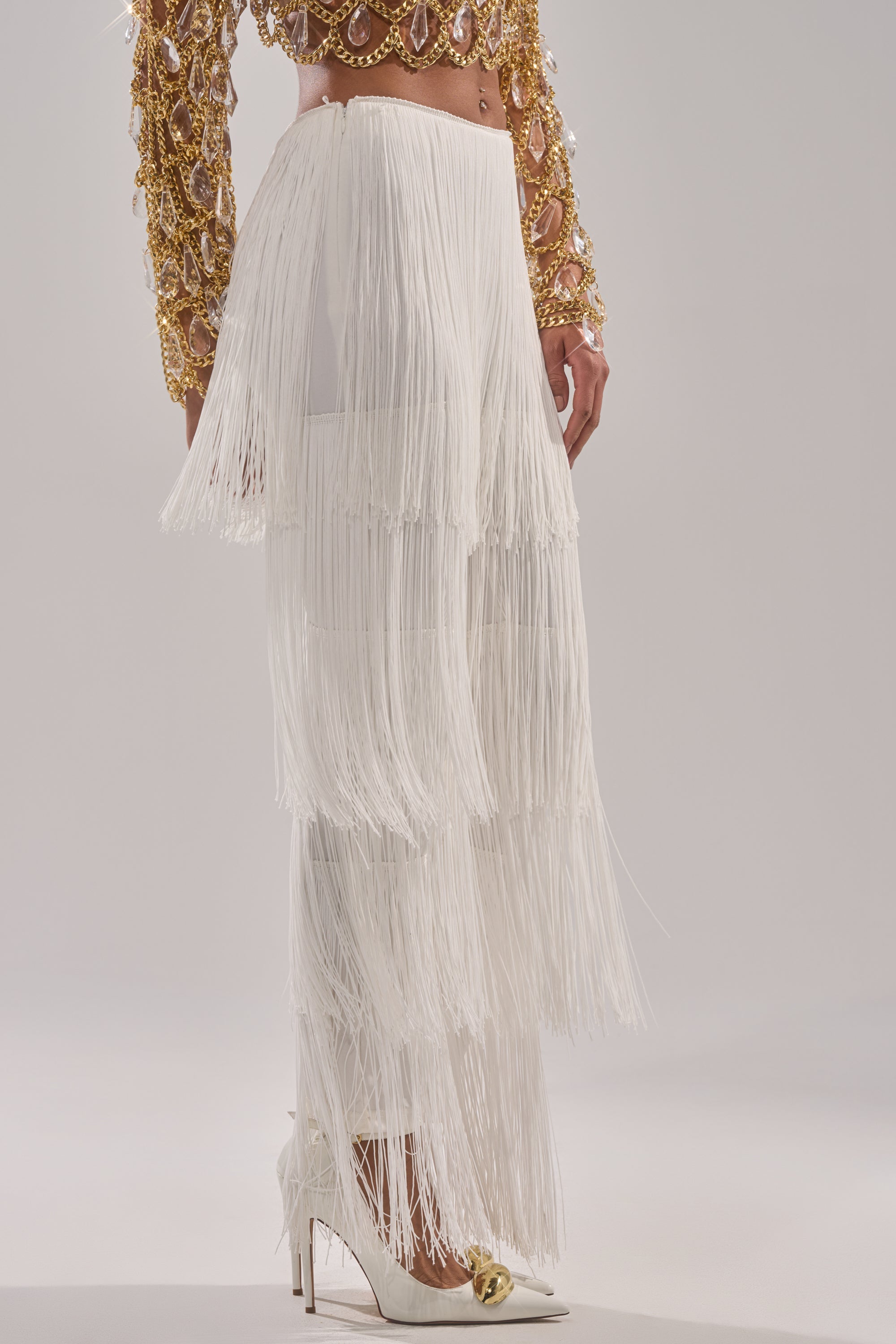 Wearing the OH SO FRINGE PANTS IN WHITE, a gold and crystal-embellished crop top, and white pointed-toe heels with gold detail, a person stands against a neutral background. 3