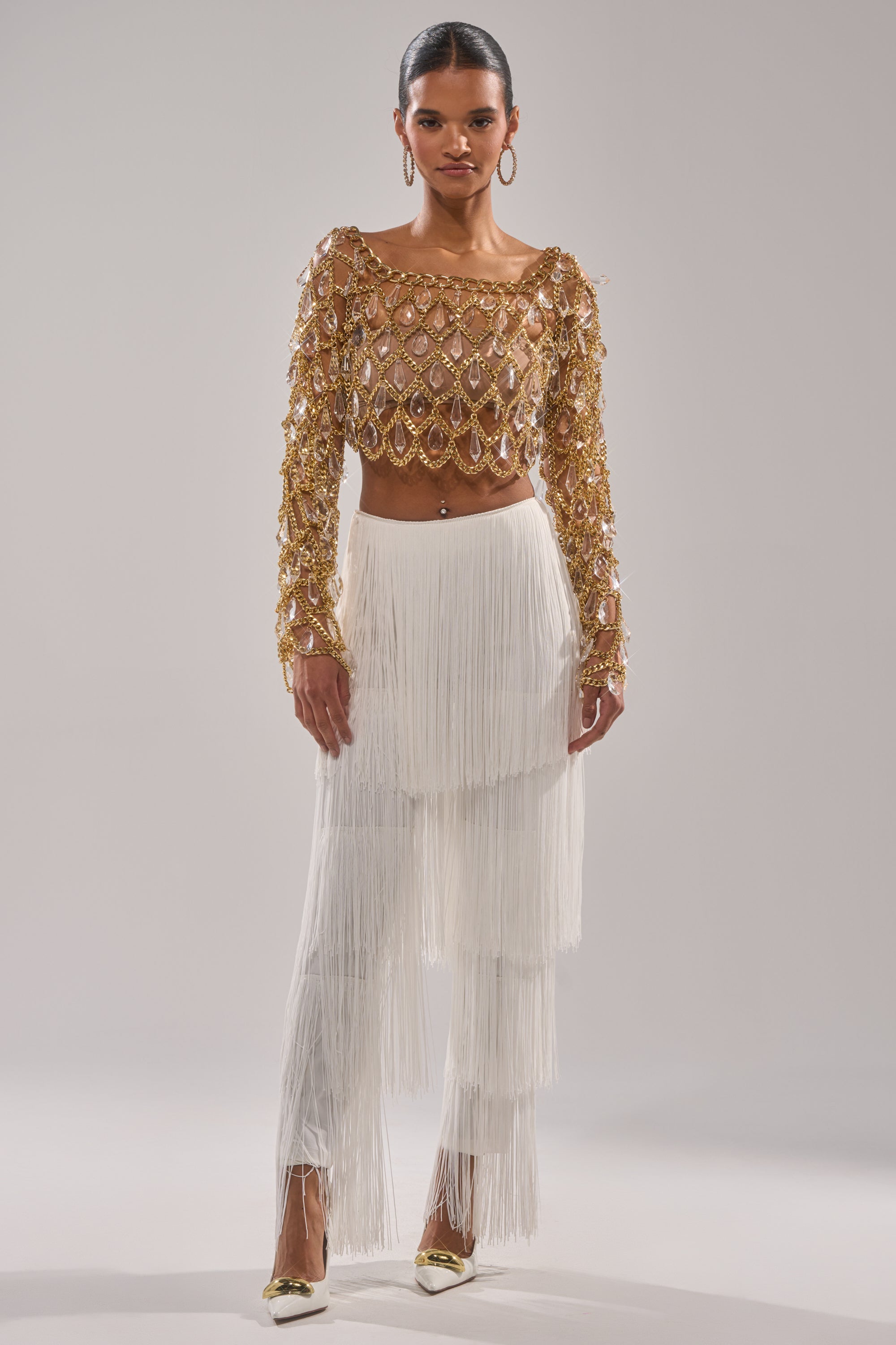 A woman faces forward in a gold, gem-encrusted crop top and the OH SO FRINGE PANTS IN WHITE, paired with pointed gold heels against a plain light gray background. 5