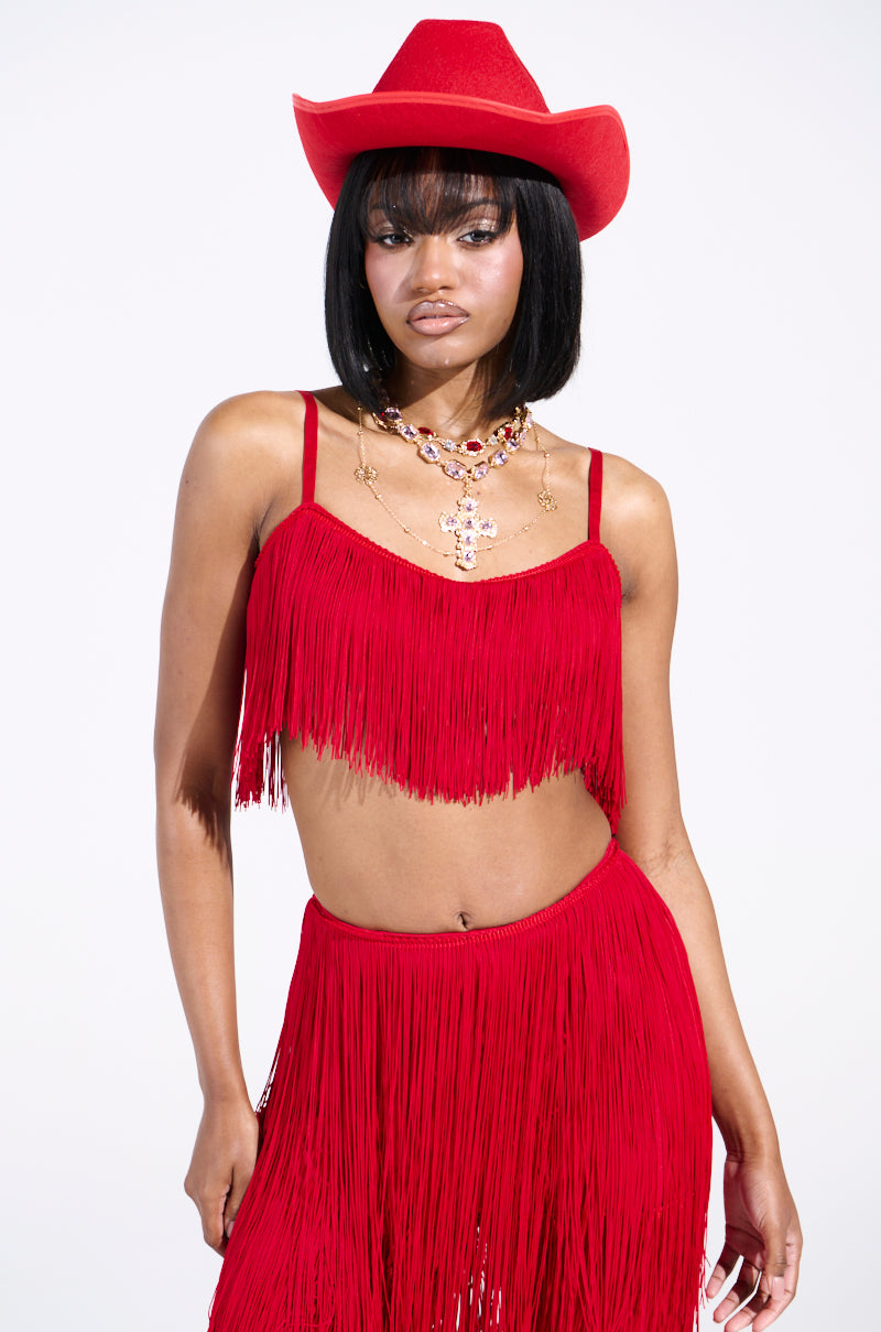 A person in the OH SO FRINGE TOP IN RED and matching cowboy hat poses confidently. They accessorize with layered necklaces and have a stylish bob haircut, set against a plain white background, emphasizing the outfit's vibrant color and eye-catching metallic zipper detail.
