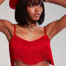 A woman confidently poses in the OH SO FRINGE TOP IN RED with a chic metallic zipper, paired with matching pants and a cowboy hat. She accessorizes with a statement gold necklace featuring a large cross, arms raised to adjust her hat against the plain background.