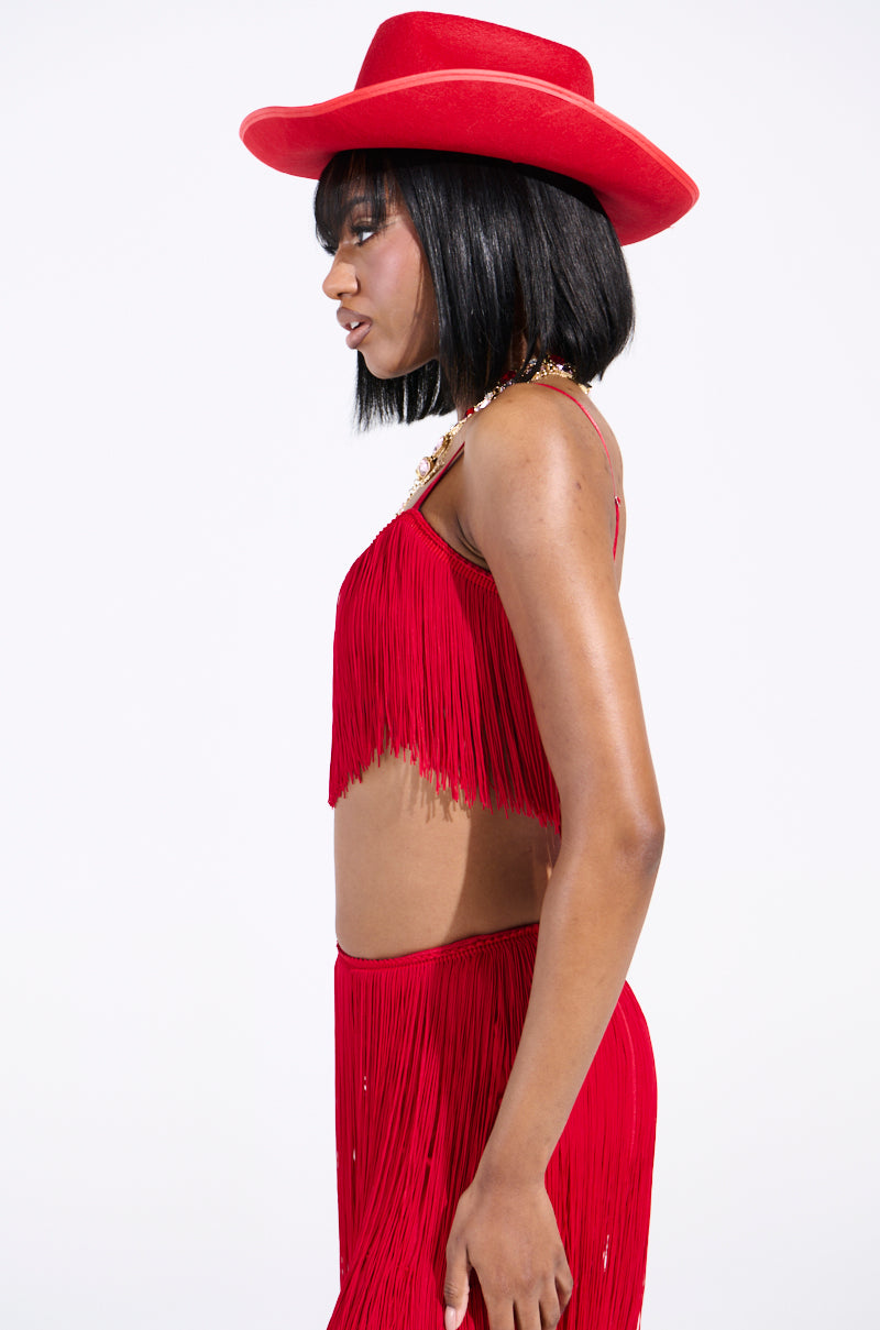 Someone wearing the OH SO FRINGE TOP IN RED and a matching wide-brimmed hat stands in profile against a plain white background. Their shoulder-length black hair frames their face as they gaze downward, exuding an air of quiet introspection.