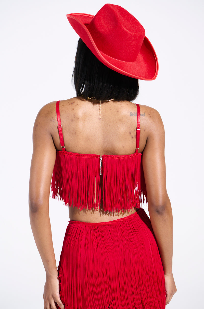 Wearing the OH SO FRINGE TOP IN RED and a matching skirt with a wide-brimmed hat, a person stands out against the white background. The vibrant ensemble is eye-catching as they face away from the camera.