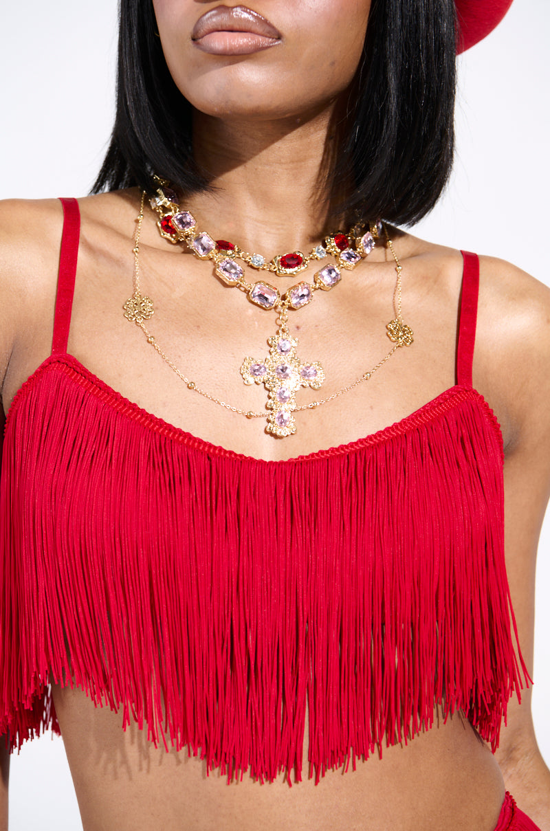 A woman wears the OH SO FRINGE TOP IN RED with a bralette silhouette and a red hat. Her layered gold necklace, adorned with large colored gemstones and a striking cross pendant, highlights her torso, while the outfit's texture is accented by a metallic zipper closure at the back.
