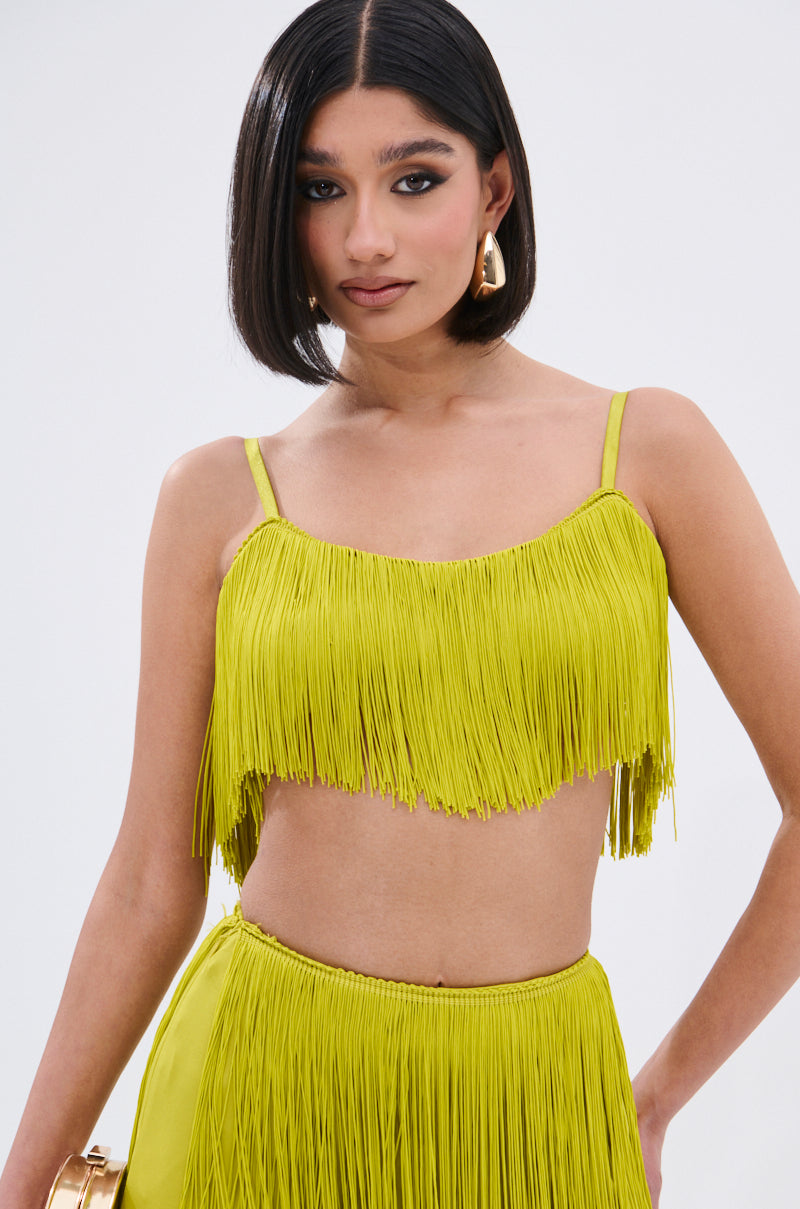 A woman with straight dark hair, gold earrings, and bold makeup wears the OH SO FRINGE TOP IN GREEN with adjustable straps and a matching skirt, posing confidently against a plain white background.