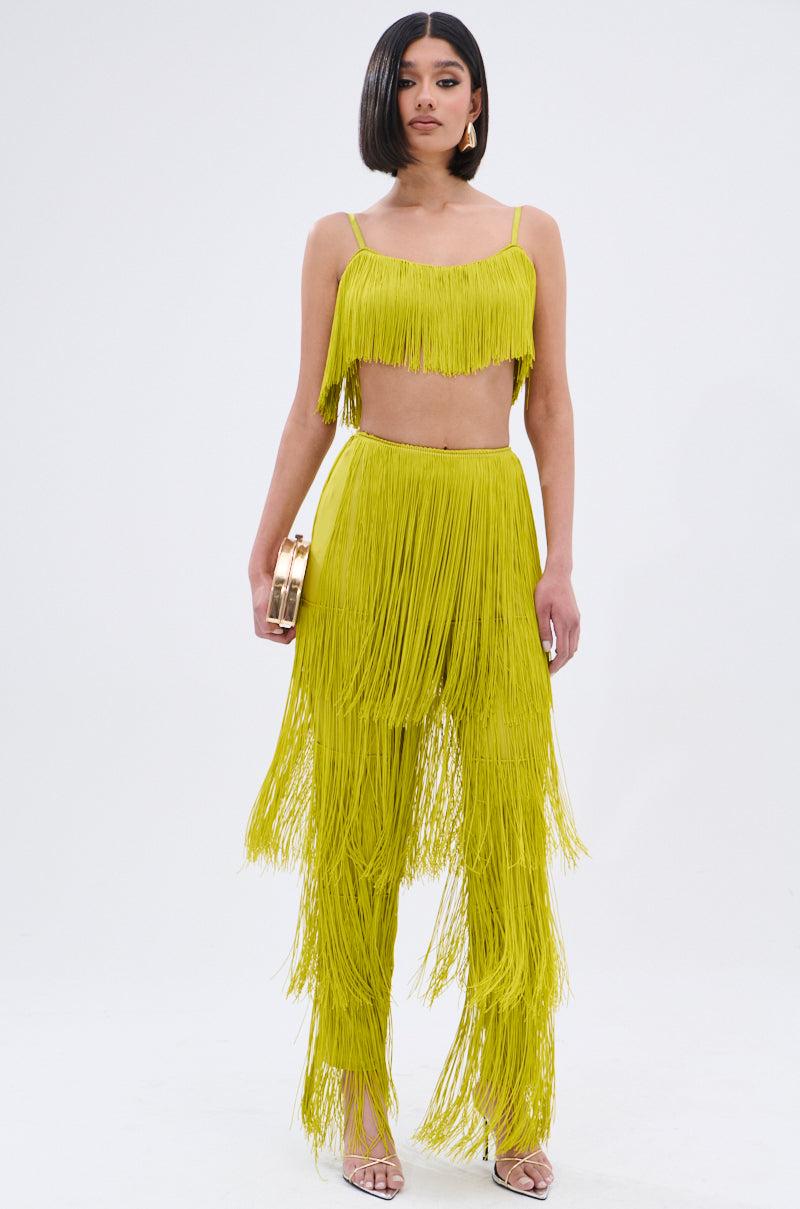 A woman stands against a white background in the OH SO FRINGE TOP IN GREEN with adjustable straps, paired with matching high-waisted fringe pants, holding a gold clutch and wearing strappy sandals.