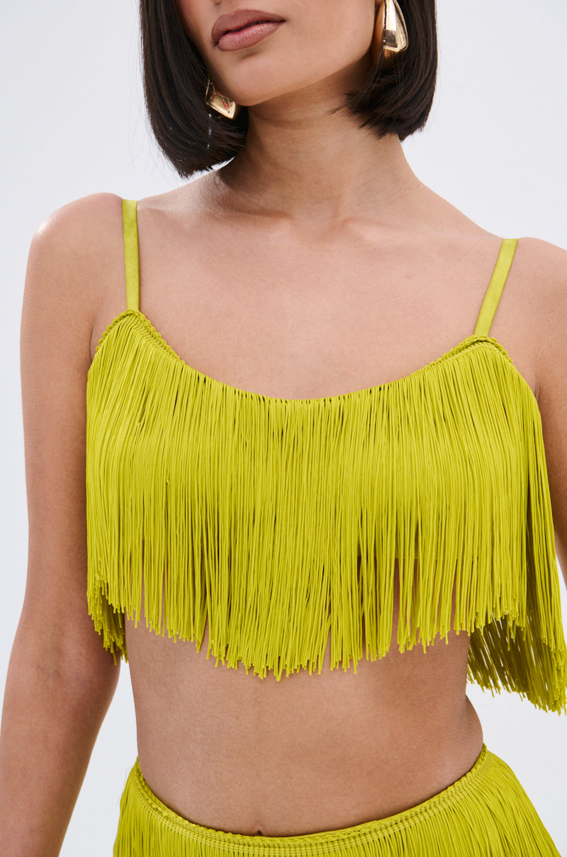 A woman with a bob haircut and large gold hoop earrings models the OH SO FRINGE TOP IN GREEN, featuring adjustable straps and fringe detail; only her torso and lower face are visible.