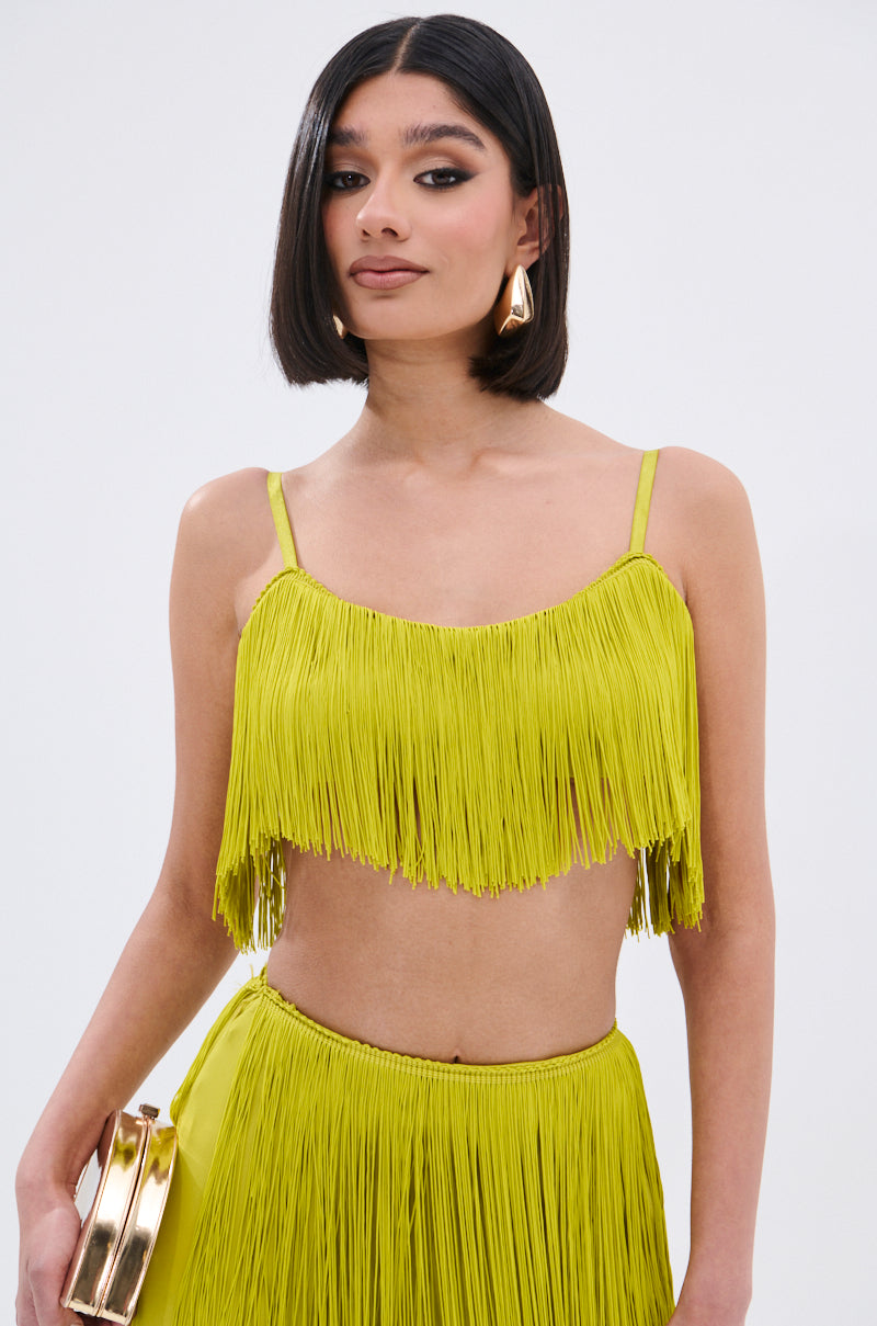 A woman with a sleek bob confidently poses against a plain white background, wearing the OH SO FRINGE TOP IN GREEN and a matching skirt with metallic zipper details. She accessorizes with a gold clutch and statement earrings.