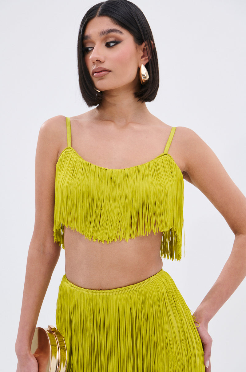 A woman with straight, shoulder-length dark hair models the OH SO FRINGE TOP IN GREEN, featuring a metallic zipper closure and fringe detail, paired with a matching skirt, gold earrings, and a clutch against a plain light background.