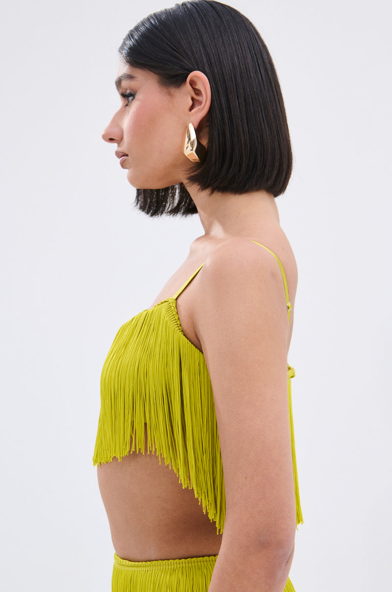A woman with straight, dark, shoulder-length hair is shown in profile wearing the OH SO FRINGE TOP IN GREEN and large gold geometric earrings against a plain light background.