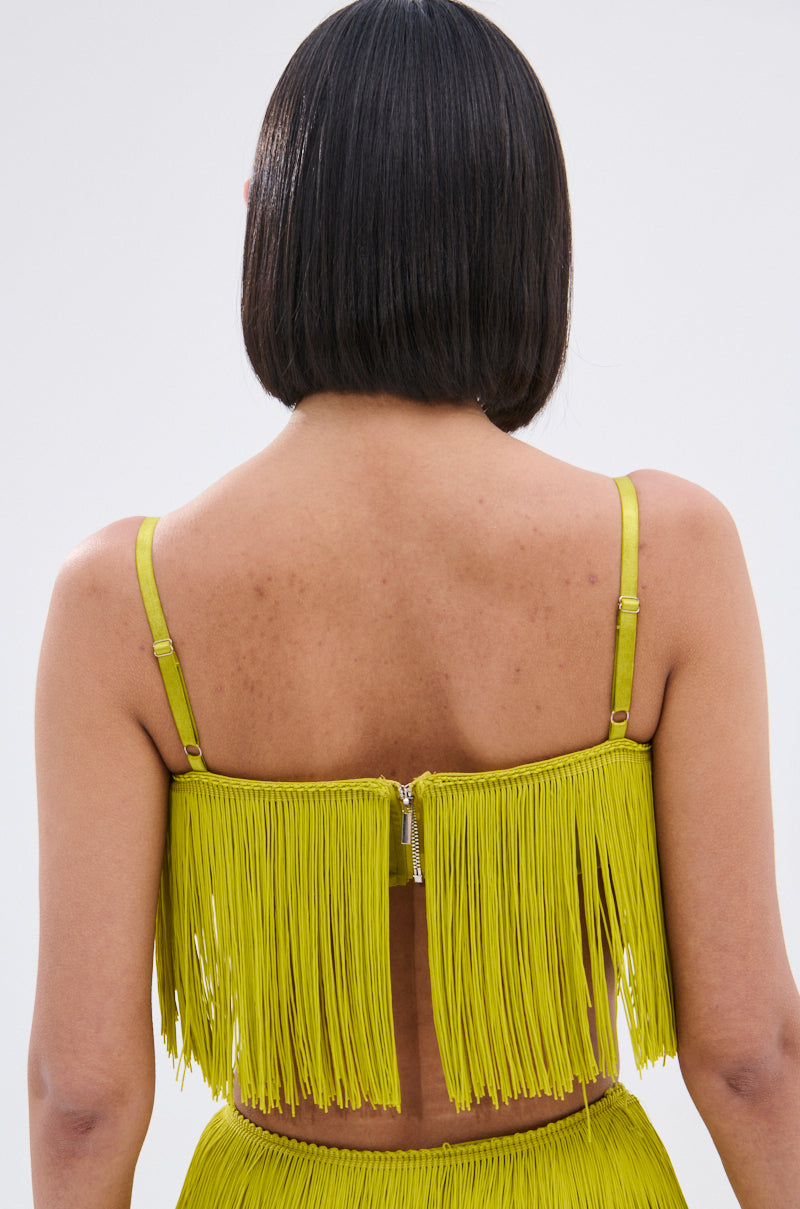 A woman with straight, shoulder-length hair is shown from the back, wearing the OH SO FRINGE TOP IN GREEN featuring adjustable straps and a metallic zipper closure against a plain, light-colored background.