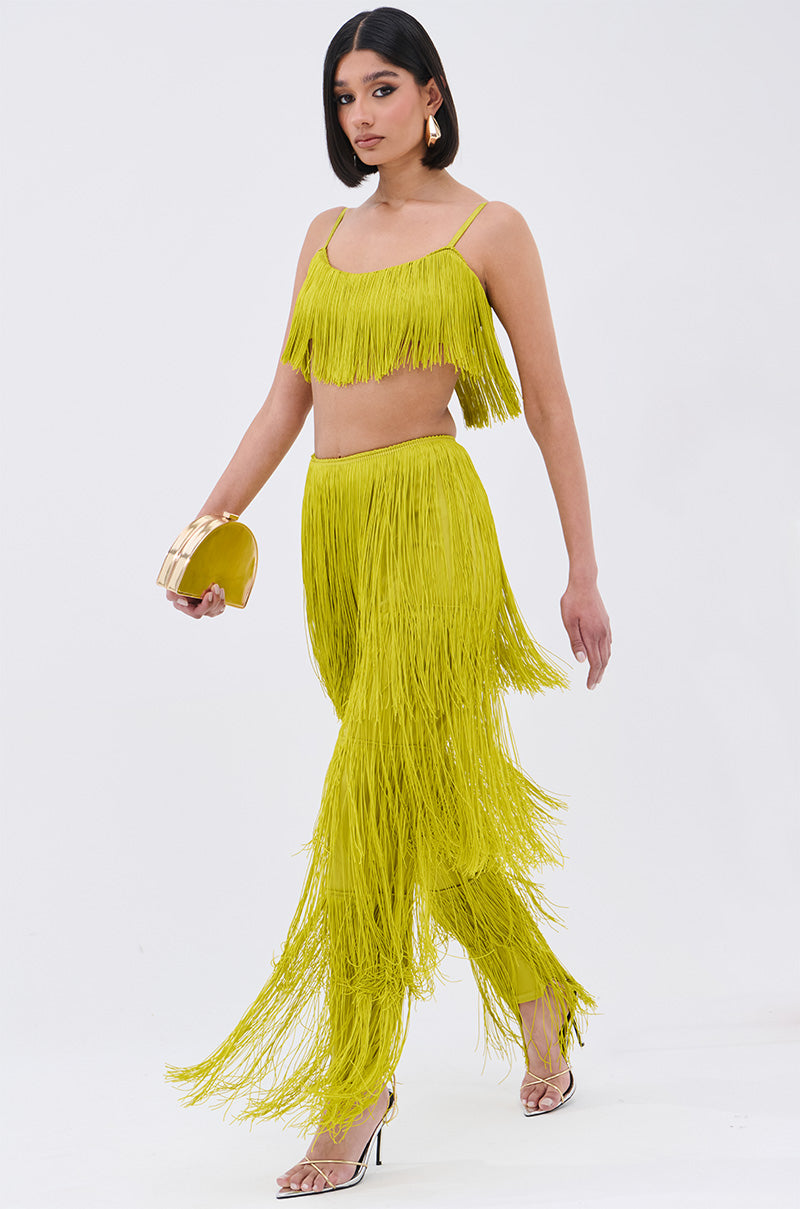 A woman wearing the OH SO FRINGE TOP IN GREEN with adjustable straps and matching high-waisted fringe pants, holding a gold clutch, and walking in clear high-heeled sandals against a plain white background.