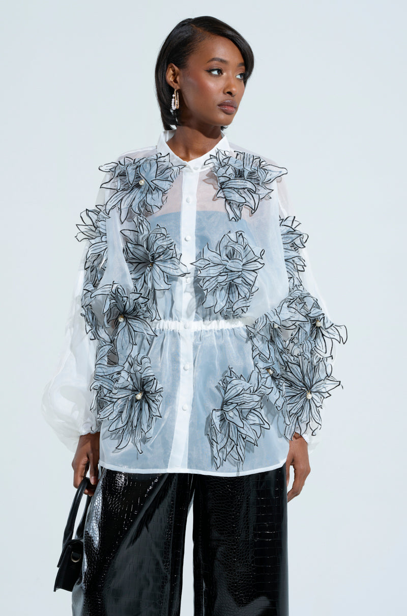Wearing the A Gentle Reminder Flower Applique Blouse made of sheer gauzy fabric and adorned with 3D flower details, a woman stylishly pairs it with black pants and holds a sleek black handbag against a light background.