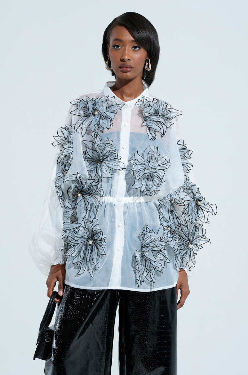 A person wearing the A GENTLE REMINDER FLOWER APPLIQUE BLOUSE, featuring a white, sheer gauzy design with prominent 3D flower appliques, pairs it with black shiny pants and holds a small handbag. Their hair is styled straight, complemented by bold earrings as they stand against a plain background.