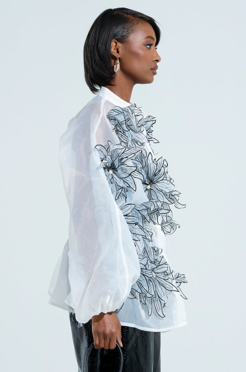 A person with short hair is wearing the A Gentle Reminder Flower Applique Blouse, which features sheer white fabric adorned with striking 3D flower appliques. They are holding a black handbag and wearing shiny black pants against a plain, light-colored background.
