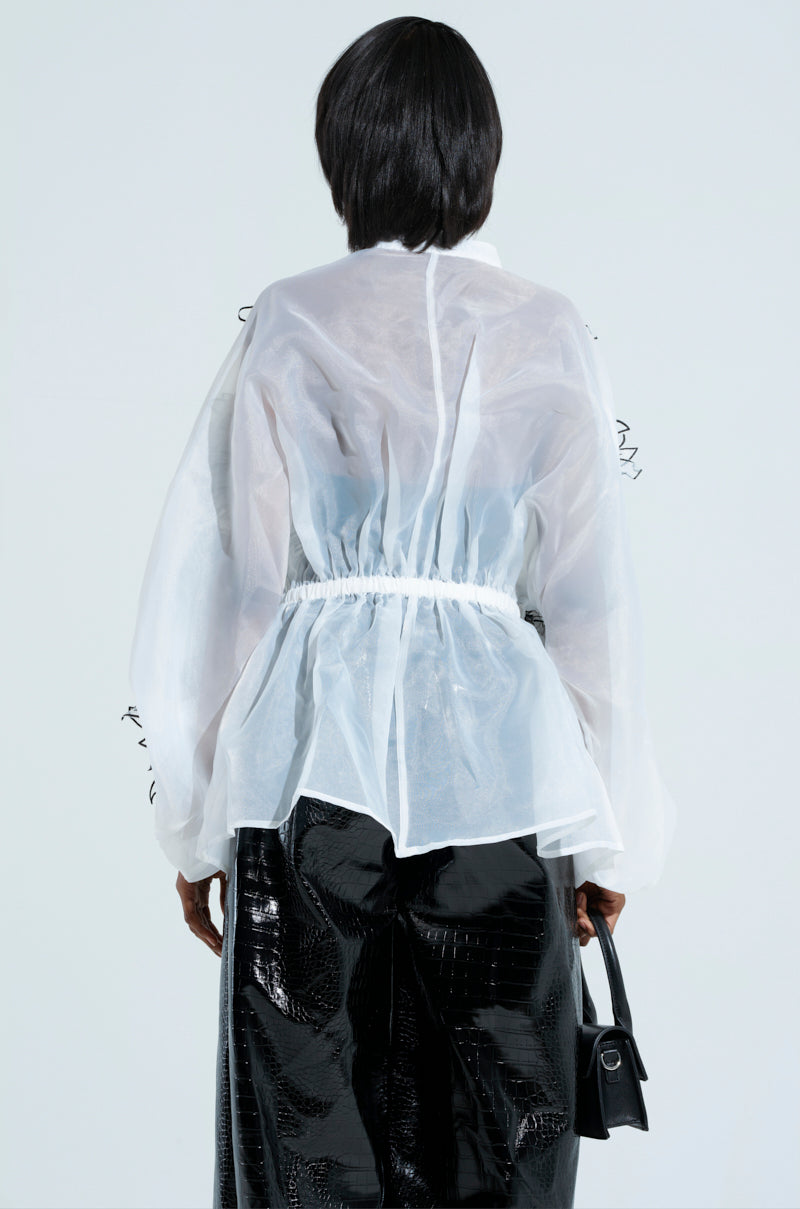 A person with short black hair is viewed from behind, dressed in the A GENTLE REMINDER FLOWER APPLIQUE BLOUSE—a translucent white long-sleeve top made from sheer gauzy fabric—and shiny black pants. They carry a small black handbag and stand before a plain white background.
