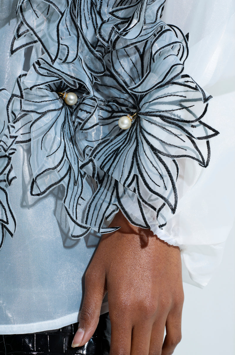 Close-up of a person's hand resting against their side, wearing the A GENTLE REMINDER FLOWER APPLIQUE BLOUSE, featuring a sheer, gauzy fabric adorned with floral patterns outlined in black and embellished with pearl-like beads. The background is a plain light color.