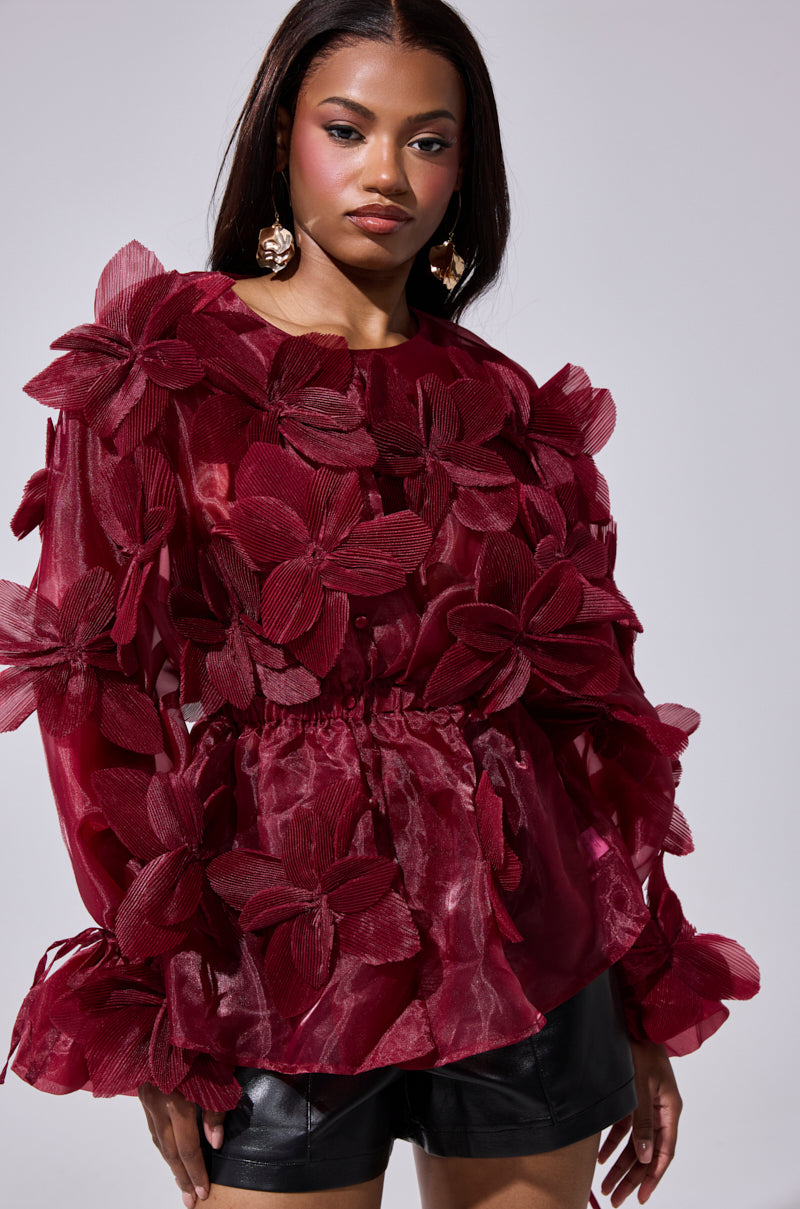 A woman wears the FLOWER POWER BLOUSE, an organza statement top in deep red with large 3D floral appliqués, paired with black leather shorts, against a plain light gray background.