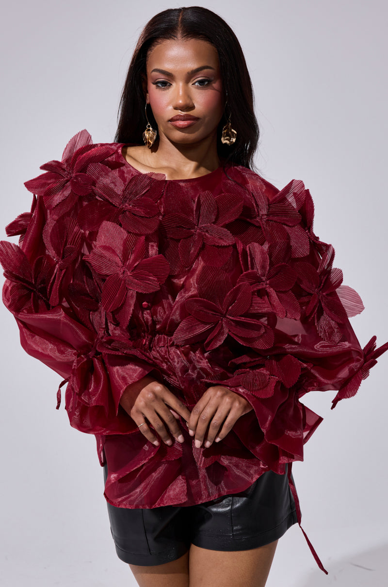 A woman with long dark hair wears the FLOWER POWER BLOUSE, featuring bold red 3D floral appliqués, paired with black leather shorts. She poses confidently against a plain, light background.