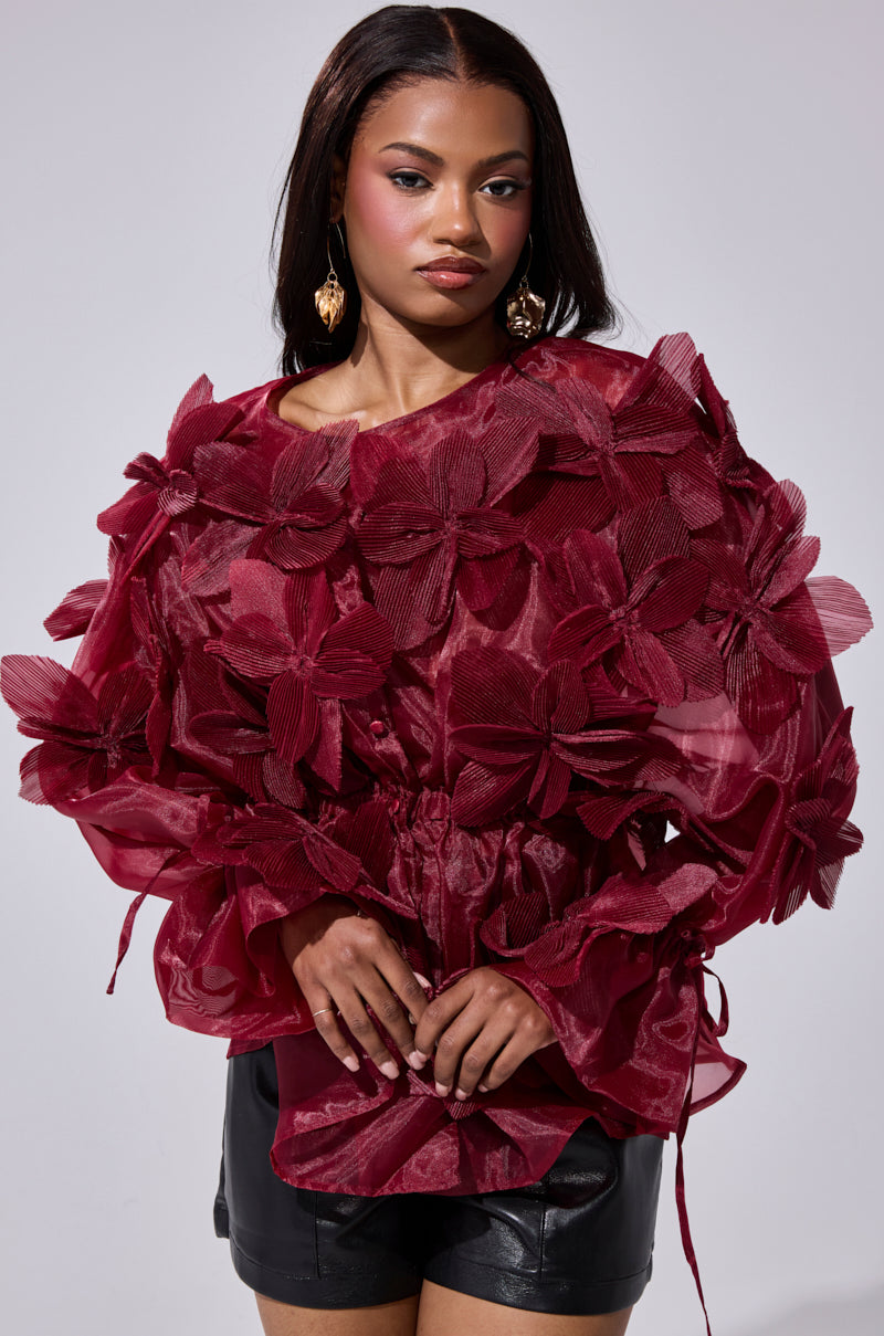 A woman stands confidently against a plain background, wearing the FLOWER POWER BLOUSE—a sheer organza top with bold floral appliqués—paired with black leather shorts, gold earrings, and long straight hair.