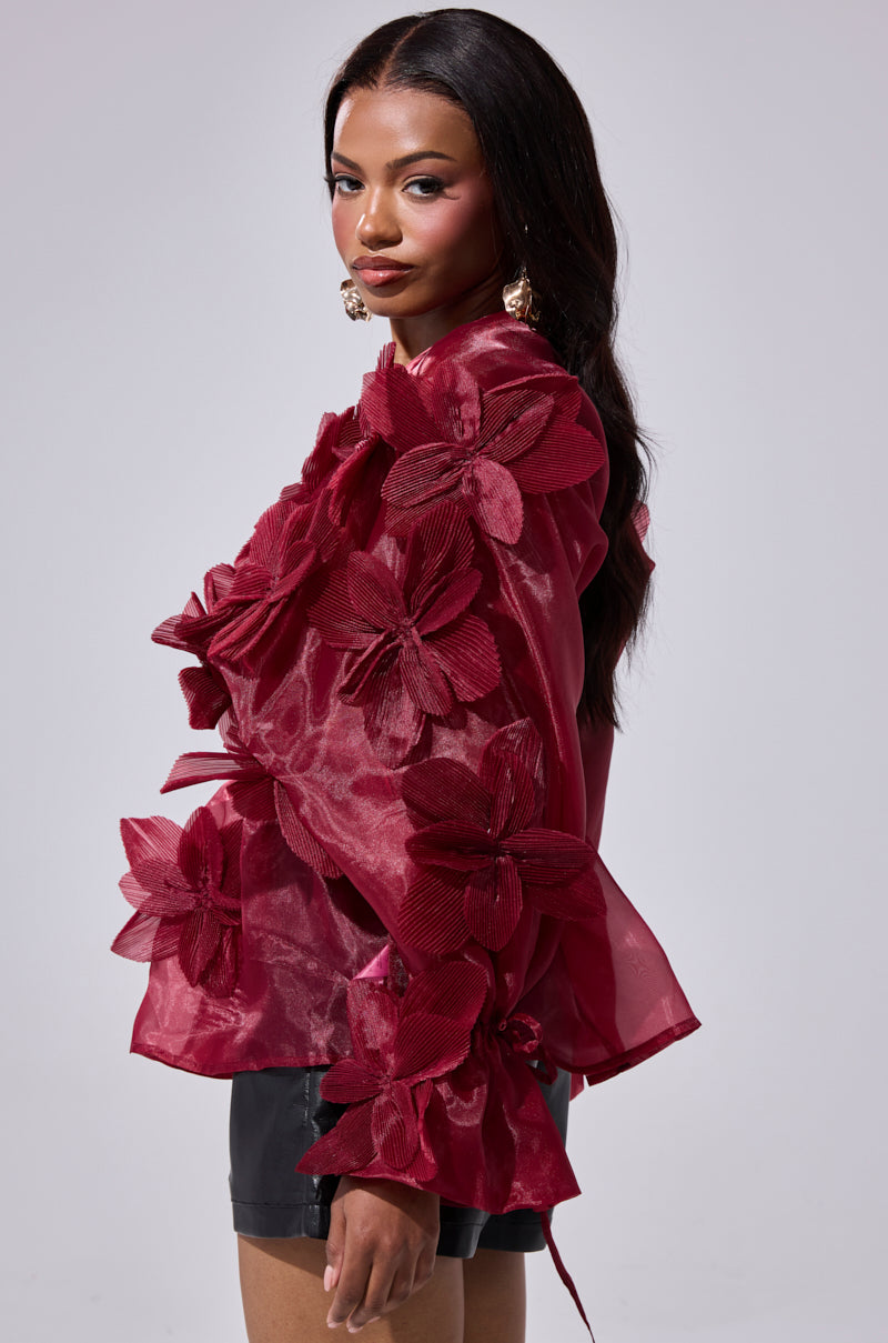 A woman with long dark hair models the FLOWER POWER BLOUSE, a deep red organza top with bold 3D floral appliqués, paired with black shorts against a plain light gray background.