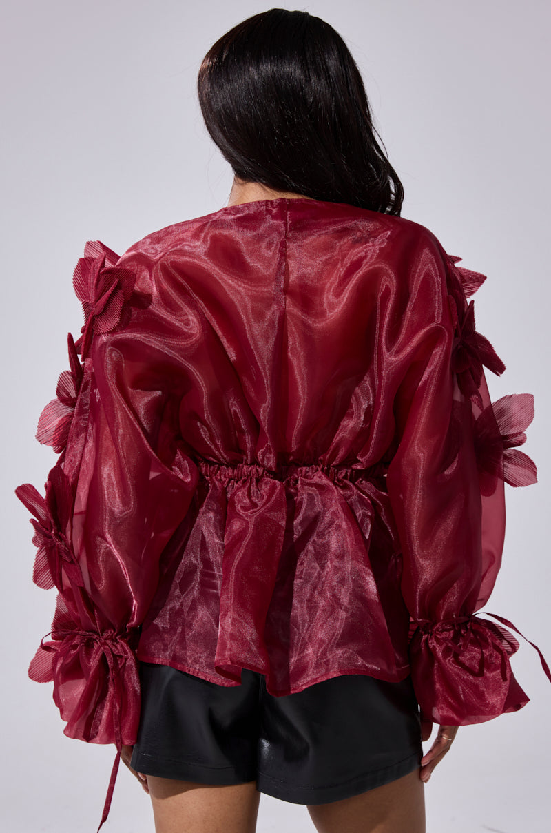 A woman with long dark hair is seen from the back, wearing the FLOWER POWER BLOUSE— a sheer deep red organza top with voluminous sleeves and striking 3D floral appliqués— paired with black shorts against a plain background.