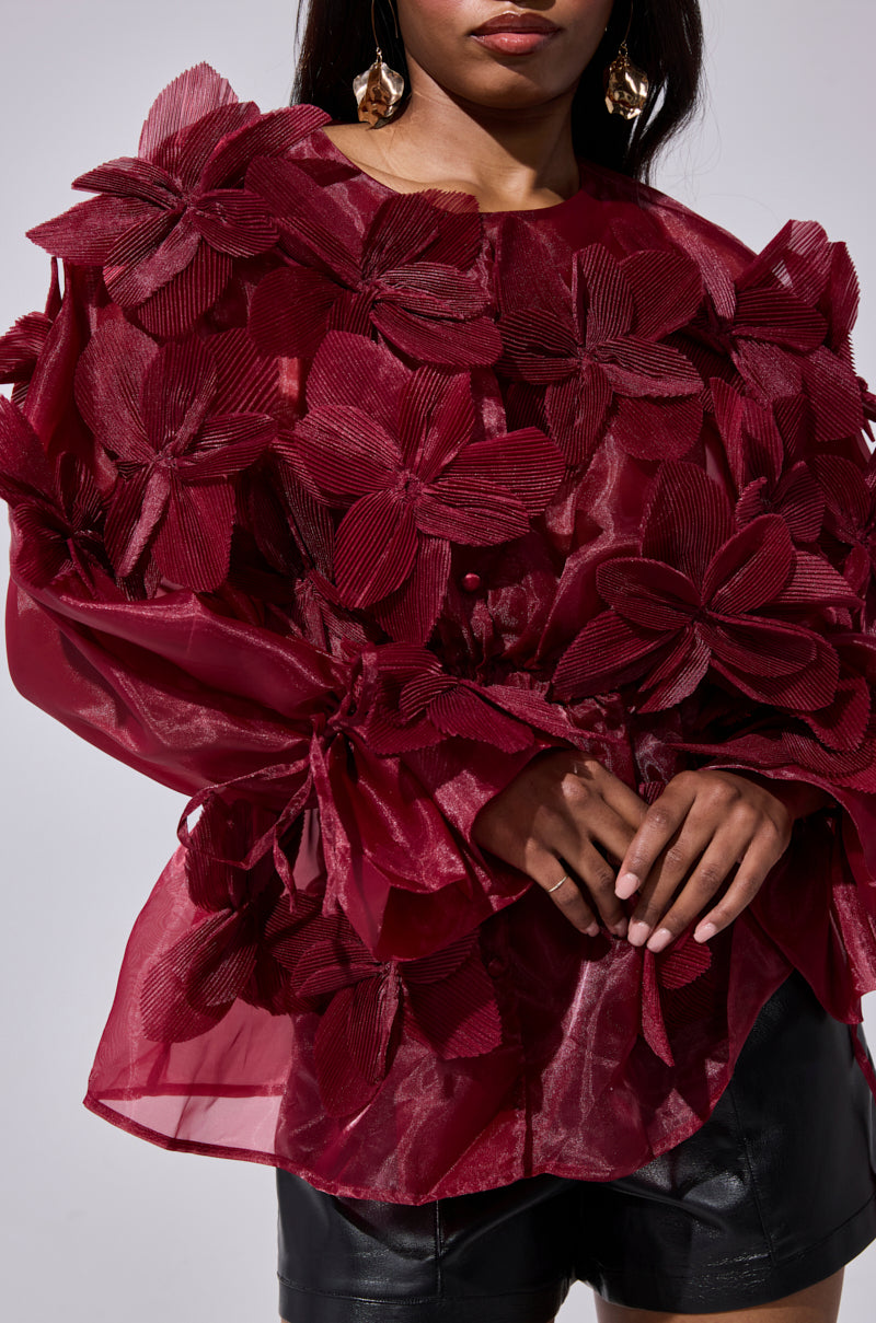 A woman models the FLOWER POWER BLOUSE—a sheer burgundy organza top with bold 3D floral appliqués—paired with black leather shorts. Her long dark hair and gold earrings frame her partially visible face.