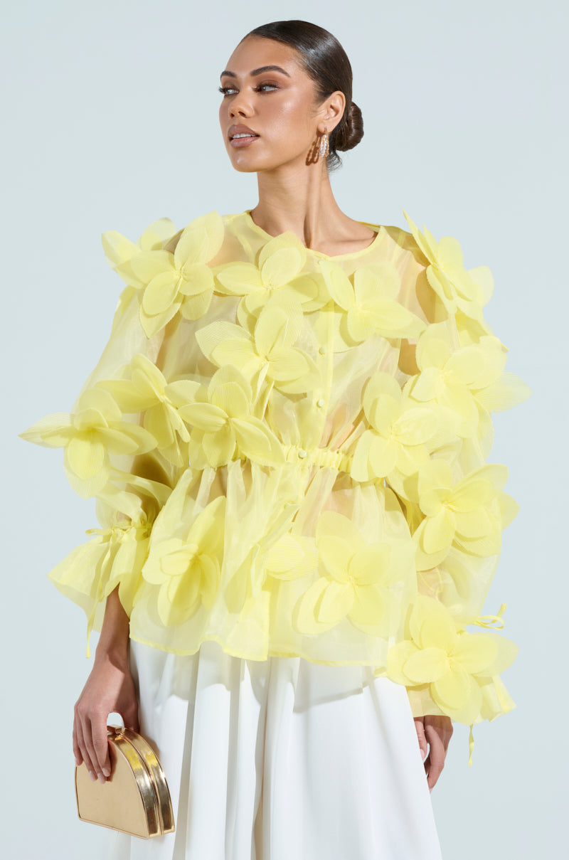 Someone wears a baby yellow Bloom Blouse with large floral appliques against a plain background. They hold a small gold clutch, wear white pants, have hoop earrings, and their hair is styled in a sleek bun.