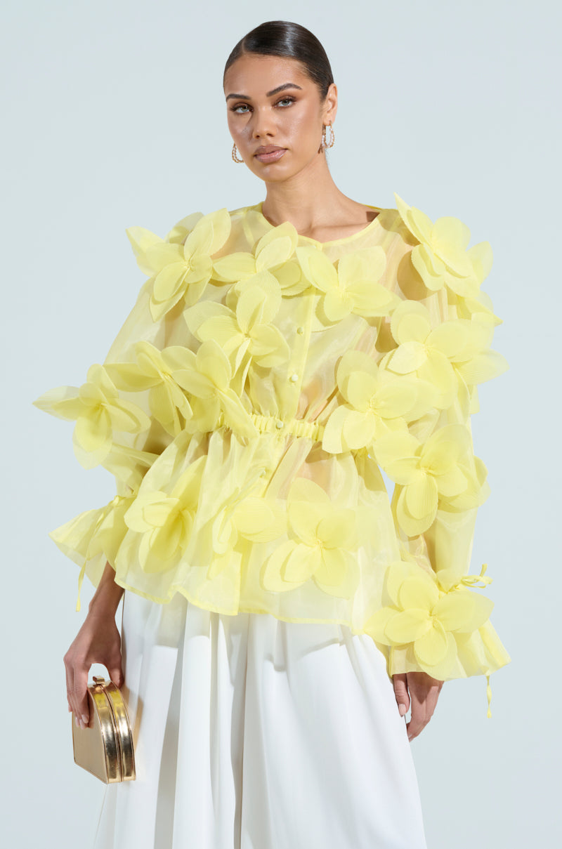A person in a BLOOM BLOUSE IN BABY YELLOW with large floral appliques stands against a plain background, holding a gold clutch and wearing white wide-legged trousers. Their hair is styled back, complemented by hoop earrings.