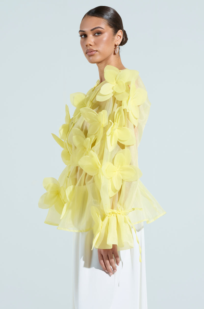 Wearing a sheer BLOOM BLOUSE IN BABY YELLOW with floral appliques, the person pairs it with white pants. They have long hair pulled back, earrings, and stand against a light blue background, looking to the side.