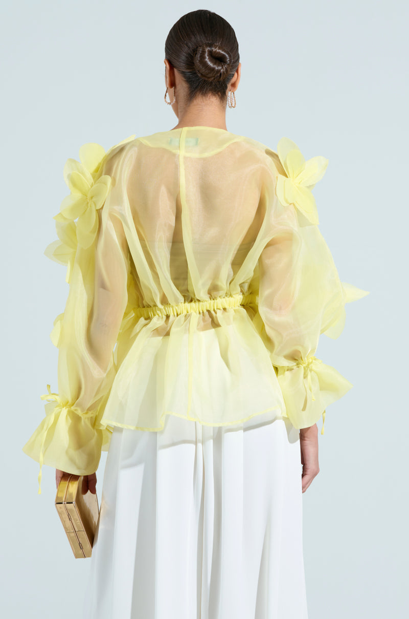 Someone with a neat bun is wearing the BLOOM BLOUSE IN BABY YELLOW, featuring large floral appliques. They're holding a small rectangular clutch and wearing white pants against a plain, light background.