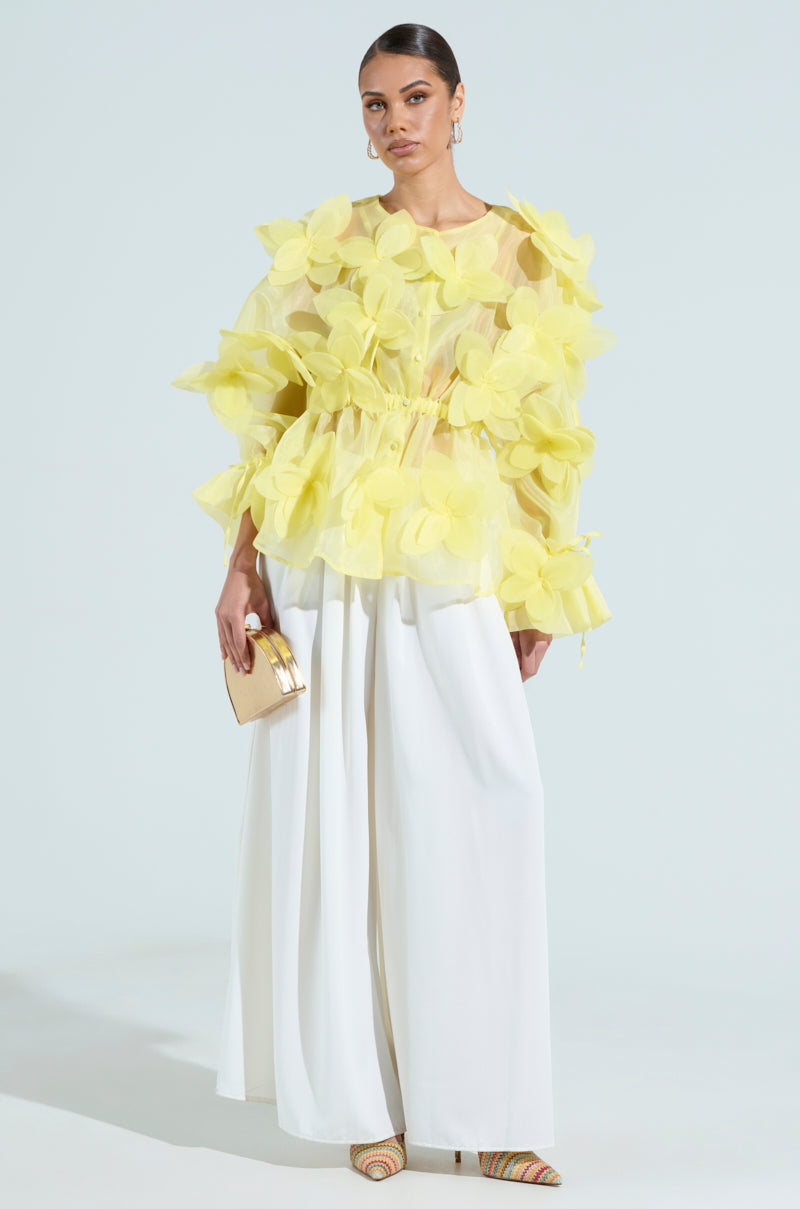 A person is in a BLOOM BLOUSE IN BABY YELLOW with floral appliques, wide-leg white pants, a small gold clutch, and sleek back hair. They wear striped heels against a light background.