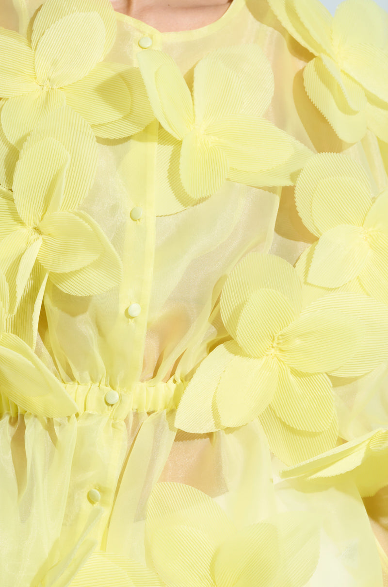 A close-up of the BLOOM BLOUSE IN BABY YELLOW reveals sheer fabric adorned with large, textured floral appliqués. The flowers have layered petals for a 3D effect, enhanced by fabric-covered buttons and a cinched waist in soft sunlight.
