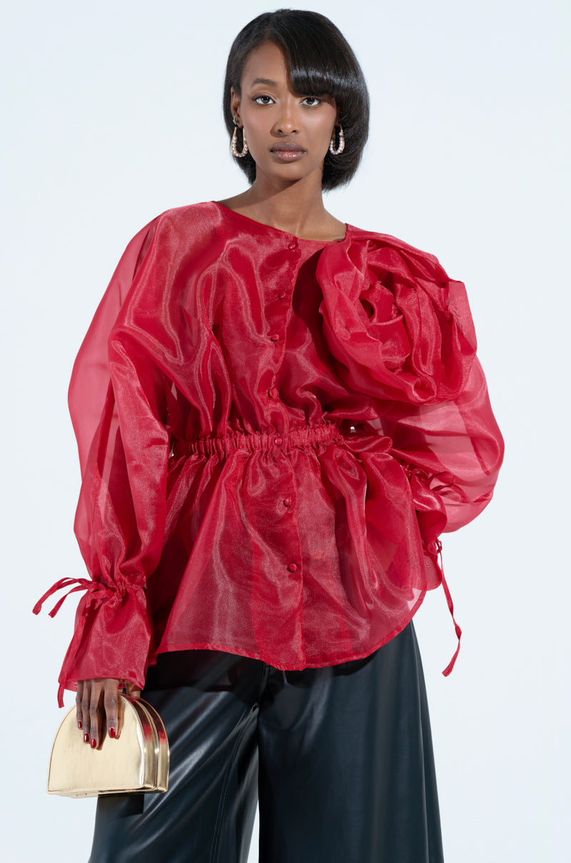 A person poses wearing a sheer red JULIA BUTTON DOWN BLOUSE with ruffled detailing, paired with black pants. They hold a small gold clutch and have hoop earrings, adding elegance with hints of peplum silhouette for flair.