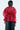 A person stands with their back turned, wearing the JULIA BUTTON DOWN BLOUSE made of sheer red chiffon, puffed sleeves, a cinched waist, and an oversized flower applique. It is stylishly paired with black pants against a plain white background.