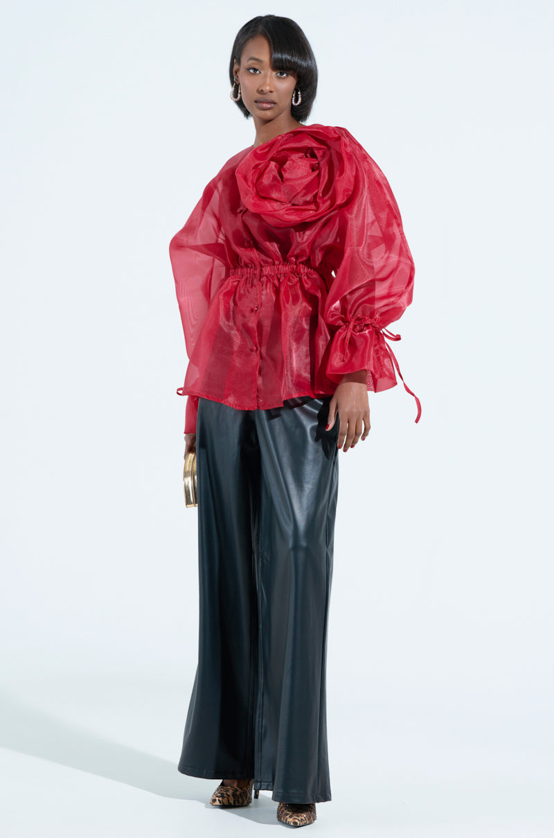 A person in a JULIA BUTTON DOWN BLOUSE, sheer and red with an oversized flower appliqué, paired with black wide-leg pants and leopard print shoes, holds a gold clutch. They stand against a white background, sporting short hair.