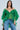 A woman with long dark hair and layered necklaces wears the AN ELEGANT EVENING ORGANZA BLOUSE in Kelly Green, featuring sheer ruffled sleeves, paired with light blue jeans. She stands against a pale background with a neutral expression.