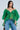 A woman wears the AN ELEGANT EVENING ORGANZA BLOUSE in sheer Kelly Green with dramatic ruffled sleeves and blue jeans. She has long dark hair, layered necklaces, and poses against a light blue background.