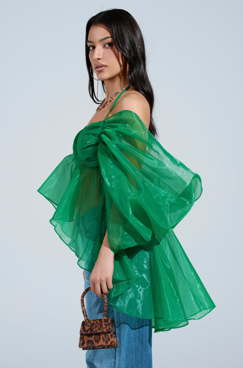 A woman poses against a plain light background wearing the AN ELEGANT EVENING ORGANZA BLOUSE in sheer, voluminous Kelly green with puffed off-the-shoulder sleeves, blue jeans, and holding a small brown animal print handbag.