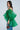 A woman poses against a plain light background wearing the AN ELEGANT EVENING ORGANZA BLOUSE in sheer, voluminous Kelly green with puffed off-the-shoulder sleeves, blue jeans, and holding a small brown animal print handbag.