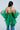 A person with long dark hair is seen from behind, wearing the AN ELEGANT EVENING ORGANZA BLOUSE in Kelly Green with voluminous sleeves and a ruffled hem, stylishly paired with blue jeans.
