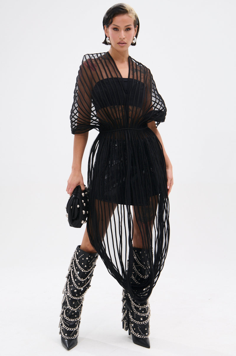 A woman poses in the RIPPLING SHEER HALTER TOP with sculptural wire-like overlays and matching earrings, carrying a small black bag and wearing boots with dangling silver chains on a plain white background.
