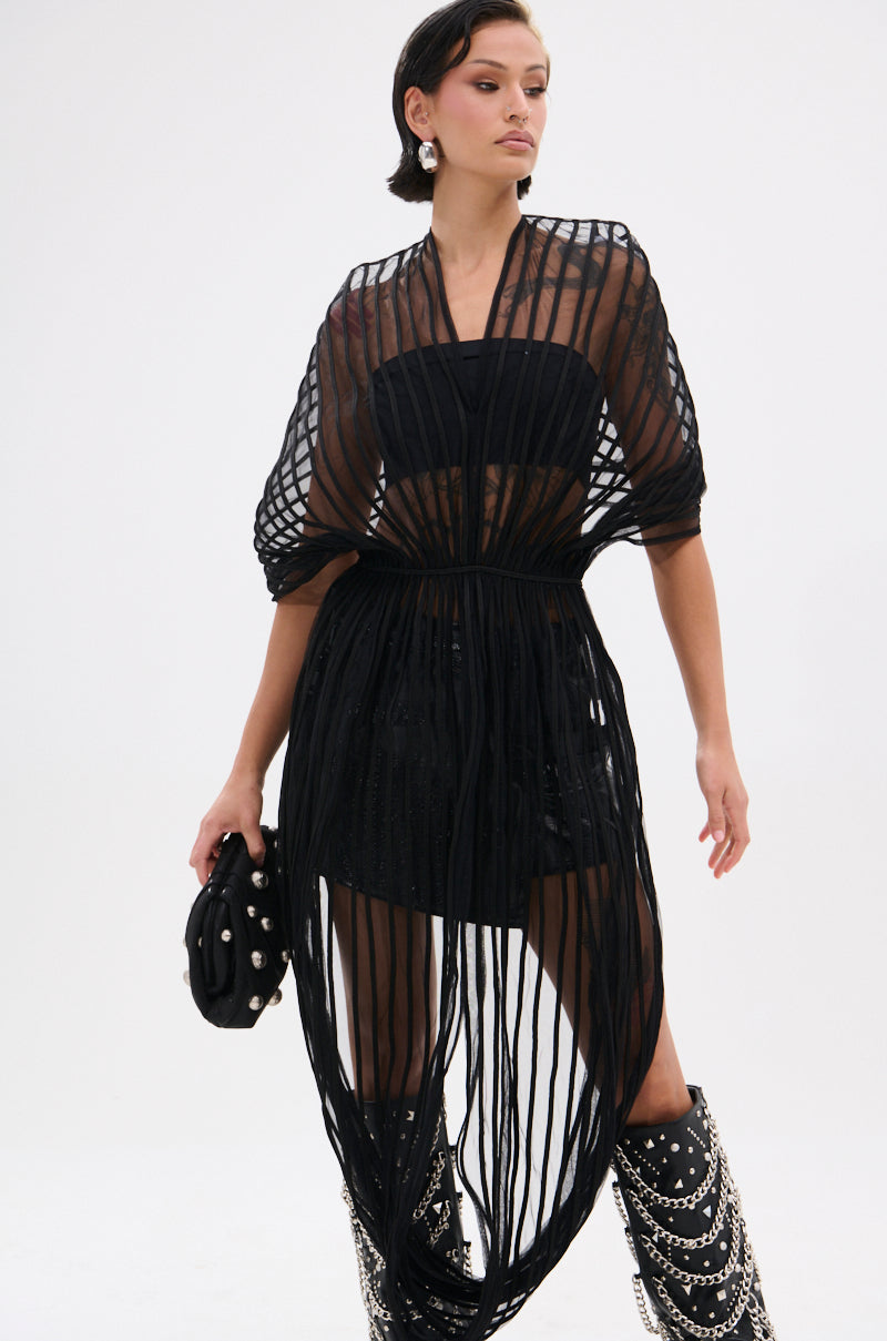 A woman poses in avant-garde fashion, wearing the RIPPLING SHEER HALTER TOP over a structured mesh bandeau and shorts, paired with a studded clutch and tall black boots embellished with silver chains.