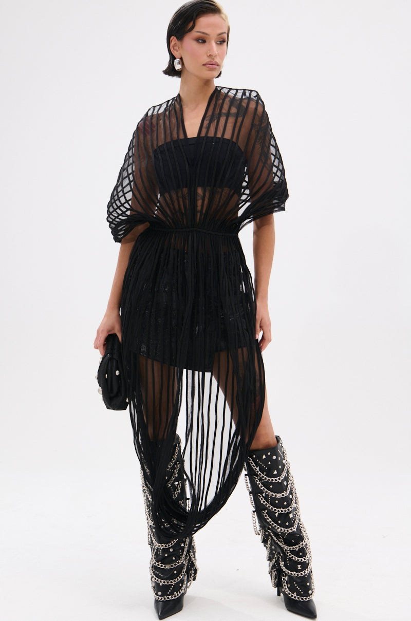 A woman poses against a white background in the RIPPLING SHEER HALTER TOP, layered over a black mini dress. She accessorizes with a small purse and black knee-high boots embellished with multiple silver chains.